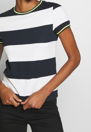 Persona que lleva una camisa de manga corta con franjas horizontales anchas en negro y blanco y ribete en verde neón en el cuello y las mangas.