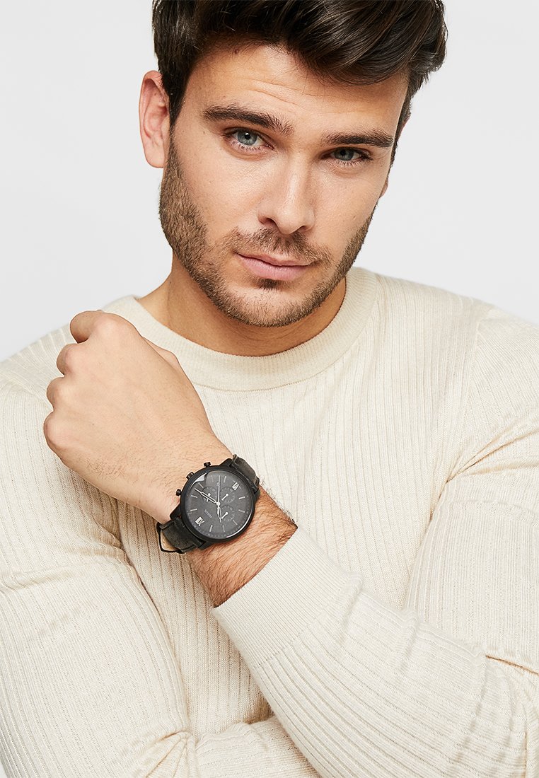 Schwarze Chronographuhr mit einem strukturierten Armband, das ein dunkles Ziffernblatt und silberne Stundenmarkierungen aufweist. Getragen an einem Handgelenk mit einem cremefarbenen, gerippten Pullover.