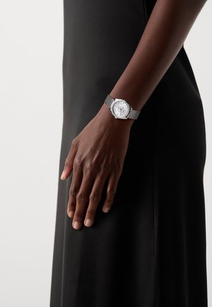 Montre-bracelet en argent avec un cadran rond, un bracelet en maille métallique, un cadran blanc et de petits marqueurs. Portée sur une main contre un fond de robe noire.