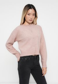 Ljust rosa kort cardigan gjord av mjuk ullblandning, med rund halsringning, ribbade mudd och knäppning framtill. Bärs över svarta jeans.