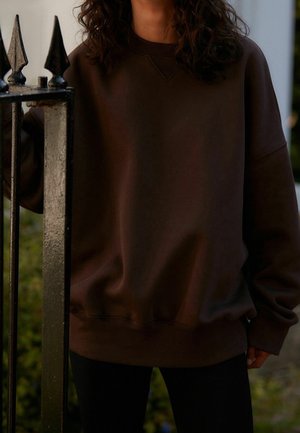 Dunkelbrauner übergroßer Sweatshirt mit rundem Halsausschnitt, gerippten Bündchen und Saum. Getragen mit schwarzen, eng anliegenden Leggings vor einem weichgezeichneten Hintergrund.