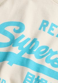 Bawełniana koszulka w kremowym kolorze z dużym turkusowym napisem w stylu vintage. Materiał ma gładką, miękką fakturę.