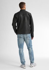 Schwarze Lederjacke mit strukturierter Oberfläche, ausgestattet mit Stehkragen und Reißverschlussärmeln, kombiniert mit hellblauen Jeans und Sneakers.