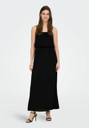 Mujer con el pelo largo y castaño, vestida con un vestido maxi negro sin mangas y sandalias negras de tacón alto, de pie y sonriendo sobre un fondo claro.