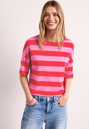 Uśmiechnięta kobieta o blond włosach, ubrana w czerwono-różową paskowaną koszulkę z krótkim rękawem i jasnoniebieskie dżinsy, ręce w kieszeniach.