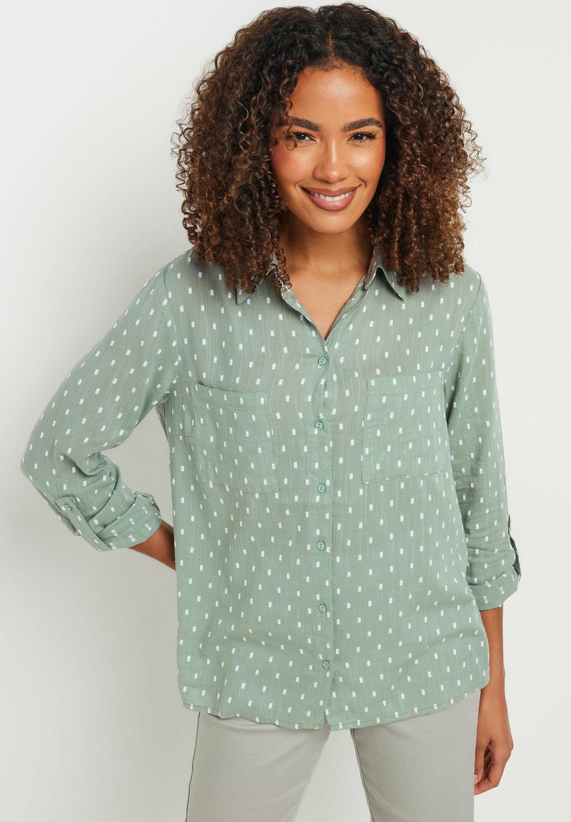 Camicia button-up verde chiaro con motivi rettangolari bianchi, due tasche frontali e maniche arrotolate. Tessuto morbido e strutturato.