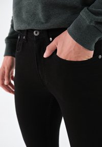 Svarta denimjeans med slim fit, med framfickor och silverfärgade knappdetaljer. Materialet har en matt yta och en slät textur.