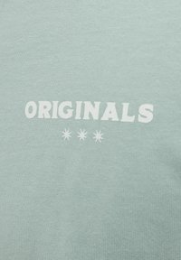 Lys grøn sweatshirt med "ORIGINALS" i fed hvid tekst og tre små stjerner nedenunder. Blødt, tekstureret stof.