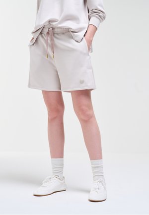 Osoba ubrana w jasnobeżowe, casualowe szorty z kieszeniami, dopasowaną bluzę z długim rękawem, białe skarpety i białe trampki na jednolitym tle.