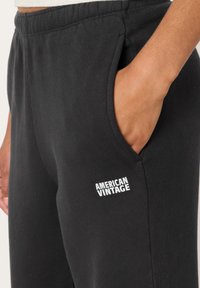 Juodi medvilniniai sportinės kelnės su šoninėmis kišenėmis, elastingu juosmeniu ir baltu „AMERICAN VINTAGE“ logotipu kairėje šlaunyje. Minkšta tekstūra.