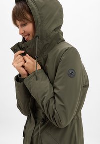 Donna che indossa un parka verde oliva con cappuccio, che sistema il colletto della giacca con entrambe le mani, sorridendo leggermente, su uno sfondo bianco semplice.
