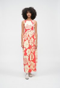 Žena s kučeravými vlasmi, oblečená v koralových maxi šatách bez rukávov s veľkými béžovými kvetinovými vzormi a v béžových topánkach, stojaca na jednofarebnom pozadí.