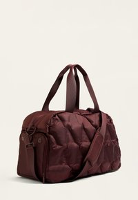 Gepolsterte bordeauxfarbene Reisetasche mit doppelten Griffen und verstellbarem Schultergurt, ausgestattet mit einer glatten Textur und einem dezenten gesteppte Design.