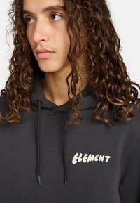 Sudadera negra hecha de un tejido suave, con capucha ajustable y un logo blanco de "ELEMENT" en el área del pecho izquierdo. Cabello texturizado visible.