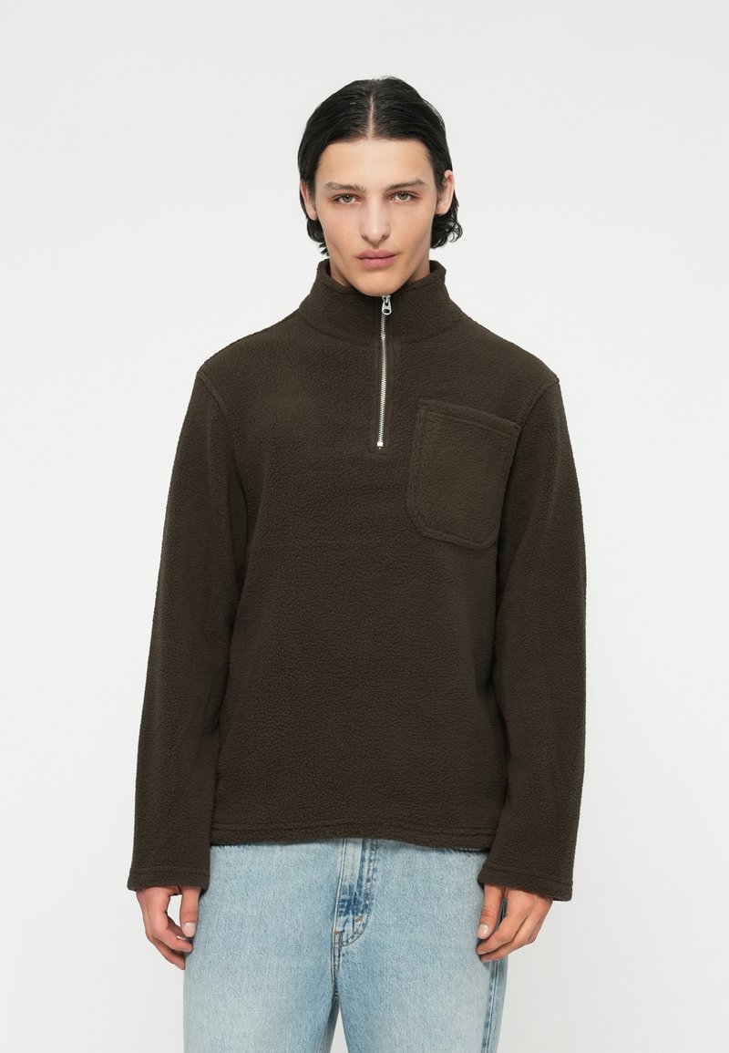 Sudadera de forro polar marrón con un alto cuello, cierre de media cremallera y un bolsillo frontal. Presenta una textura suave y mangas largas.