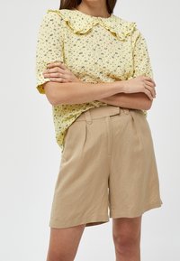 Blusa gialla a fiori con colletto arricciato, abbinata a pantaloni corti plissettati beige. Il tessuto sembra strutturato, leggero e informale.
