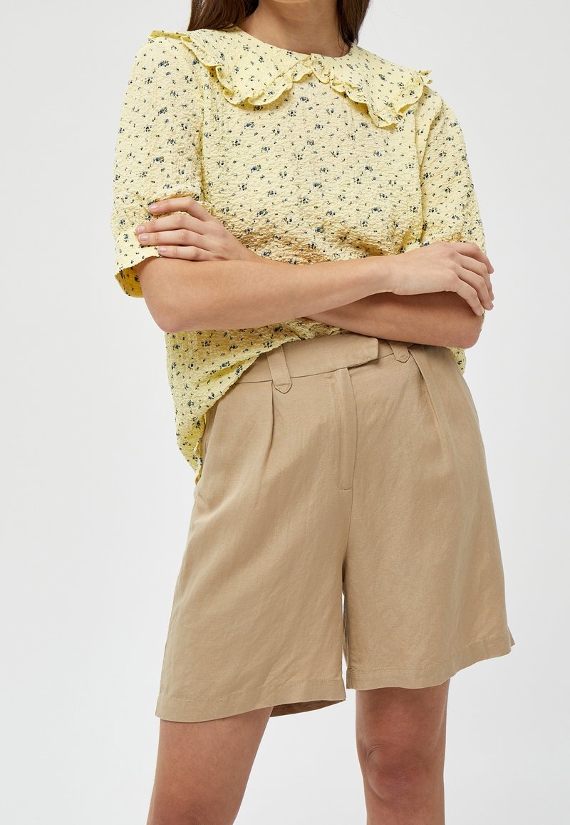 Blusa gialla a fiori con colletto arricciato, abbinata a pantaloni corti plissettati beige. Il tessuto sembra strutturato, leggero e informale.