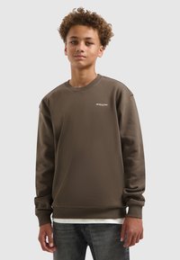 Bruine sweatshirt met geribbelde manchetten en zoom, met een wit logo op de borst. Model draagt het over een wit shirt en grijze jeans.