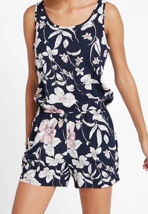 Marineblå ærmeløs romper med hvidt og lyserødt blomsterprint, elastisk talje og løs pasform på en person med hænderne i lommerne.