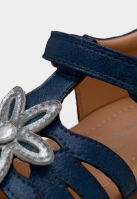 Μπλε δερμάτινο λουράκι σανδάλι με ασημένια διακόσμηση λουλουδιού και ταμπά ραμμένη εσωτερική σόλα ορατή στο φόντο.