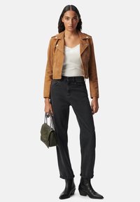 Mujer con chaqueta de ante marrón corta, blusa blanca con encaje en el borde, jeans negros de cintura alta, botas negras, sosteniendo un bolso verde acolchado con cadenas.
