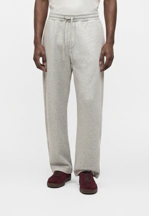 Homme portant un pantalon de survêtement gris clair avec cordon et des chaussures bordeaux à lacets, debout devant un fond blanc.