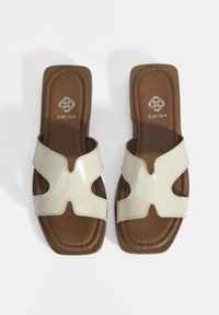 Crème kleurige gepatenteerde slider sandalen met een open teen ontwerp, voorzien van gebogen uitsparingen en een bruine gevoerde zool. Merklogo op de binnenzool.