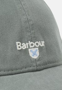 Boné de algodão verde com aba curvada, apresentando o nome da marca "Barbour" bordado e um elemento de design azul claro na parte da frente.