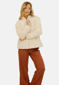 Veste en polaire écrue avec col montant et fermeture éclair, associée à un pantalon large côtelé couleur rouille. Texture douce, coupe oversize.