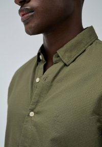 Camisa de botones verde oliva con un patrón texturizado, con cuello y botones color beige; detalle sutil del logo en el pecho.