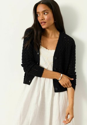 Woman with long dark hair wearing a black textured cardigan over a white dress, adjusting cardigan sleeve with a silver bracelet on left wrist.