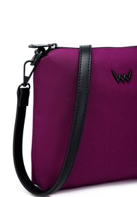 Fialová nylonová taška s černým koženým popruhem a zipem, se znakem na boku. Hladký povrch, obdélníkový tvar.