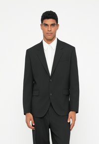 Chaqueta negra con solapas muesca, dos botones y bolsillos laterales, llevada sobre una camisa blanca. Tejido suave con un corte ajustado.