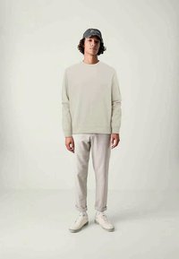 Lys grå genser med rund hals, lyse fargede skreddersydde bukser, og hvite joggesko. En mørk caps med logo fremhever det uformelle antrekket.