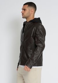 Mørkebrun læderjakke med kviltede skulderdetaljer og lynlås foran. Har en sort hætteforet indvendig samt sidelommer.