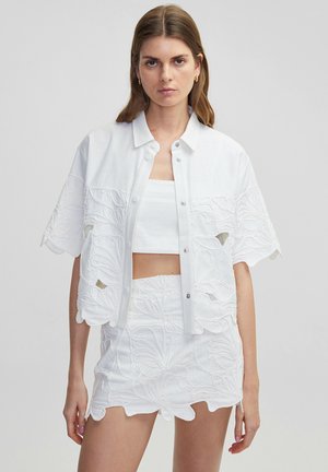 Chaqueta blanca recortada con bordados florales y botones a presión, combinada con una blusa y una falda a juego con bordes festoneados.