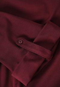 Dunkelrotes Langarmhemd mit einem mit Knöpfen versehenen Ärmelriegel, das die Stoffstruktur und Nähte zeigt.