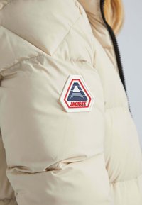 Doudoune beige avec un design matelassé, comportant un patch hexagonal proéminent avec l'inscription "JACKIT" en bleu et rouge sur la manche gauche.