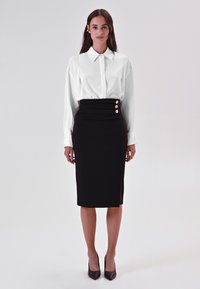 Blusa bianca a maniche lunghe con colletto, abbinata a una gonna a tubino nera fino al ginocchio con vita arricciata e tre dettagli con bottoni dorati.