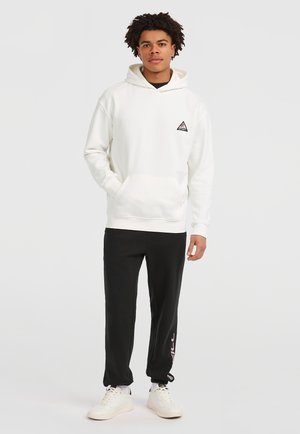 Young man standing wearing a white hoodie with a small triangle logo and black sweatpants with white sneakers on a plain background.