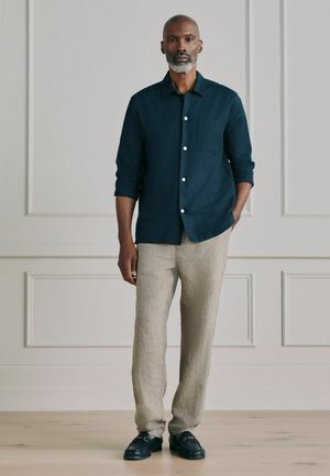 Mand med gråt skæg iført en mørkeblå skjorte med knapper, beige bukser og sorte loafers, stående op ad en væg med paneler.