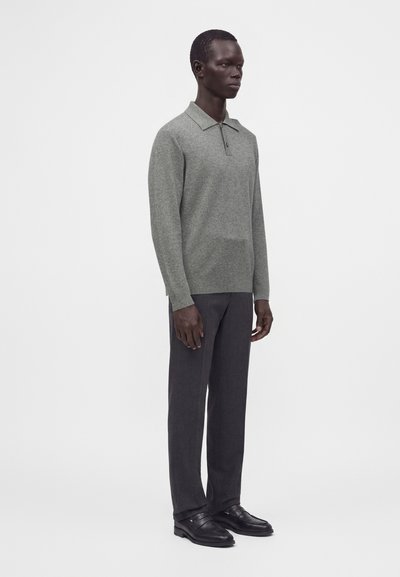 Homme portant un polo gris à manches longues, un pantalon gris foncé et des chaussures habillées noires, debout devant un fond blanc uni.