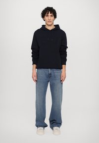 Felpa blu navy con tasca frontale e cordino, abbinata a jeans a gamba larga di un blu chiaro e sneakers bianche, su uno sfondo semplice.