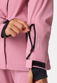 Veste rose avec des accents noirs, dotée d'une poche zippée sur la manche, de poignets ajustables et d'un détail imprimé "SKYT 5000".