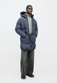 Μπουφάν puffer σε ναυτικό μπλε με κουκούλα, κλείσιμο με φερμουάρ, τσέπες στο μπροστινό μέρος και υφή, φορεμένο πάνω από γκρι ζιβάγκο και φαρδιά σκουρόχρωμα παντελόνια.