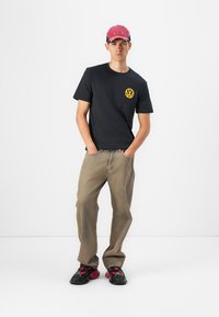 Sort t-shirt med gul Volkswagen-logo, beige baggy bukser og sort/røde sneakers. Rød kasket med hvide detaljer fuldender outfittet.
