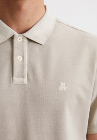 Beżowa koszulka polo z teksturowanej tkaniny, z klasycznym kołnierzykiem, dwoma guzikami i małym białym haftowanym logo na piersi.