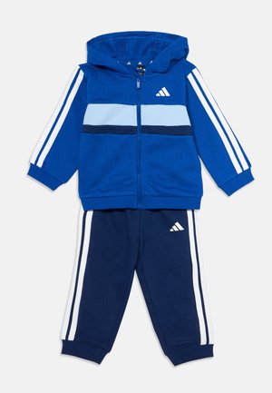 Survêtement bleu et marine pour enfants avec veste à capuche zippée et pantalon, avec des bandes blanches sur les manches et les jambes et un logo blanc de la marque sur la poitrine et la jambe.
