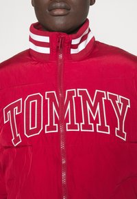 Röd vadderad jacka med hög krage och vita ränder samt en framträdande "TOMMY"-logotyp. Slät textur och dragkedja fram.
