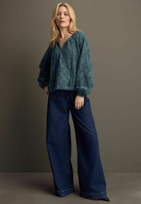 Blusa ricamata color teal con maniche a palloncino, abbinata a pantaloni in denim scuri a gamba larga a vita alta e calzature nere con punta.